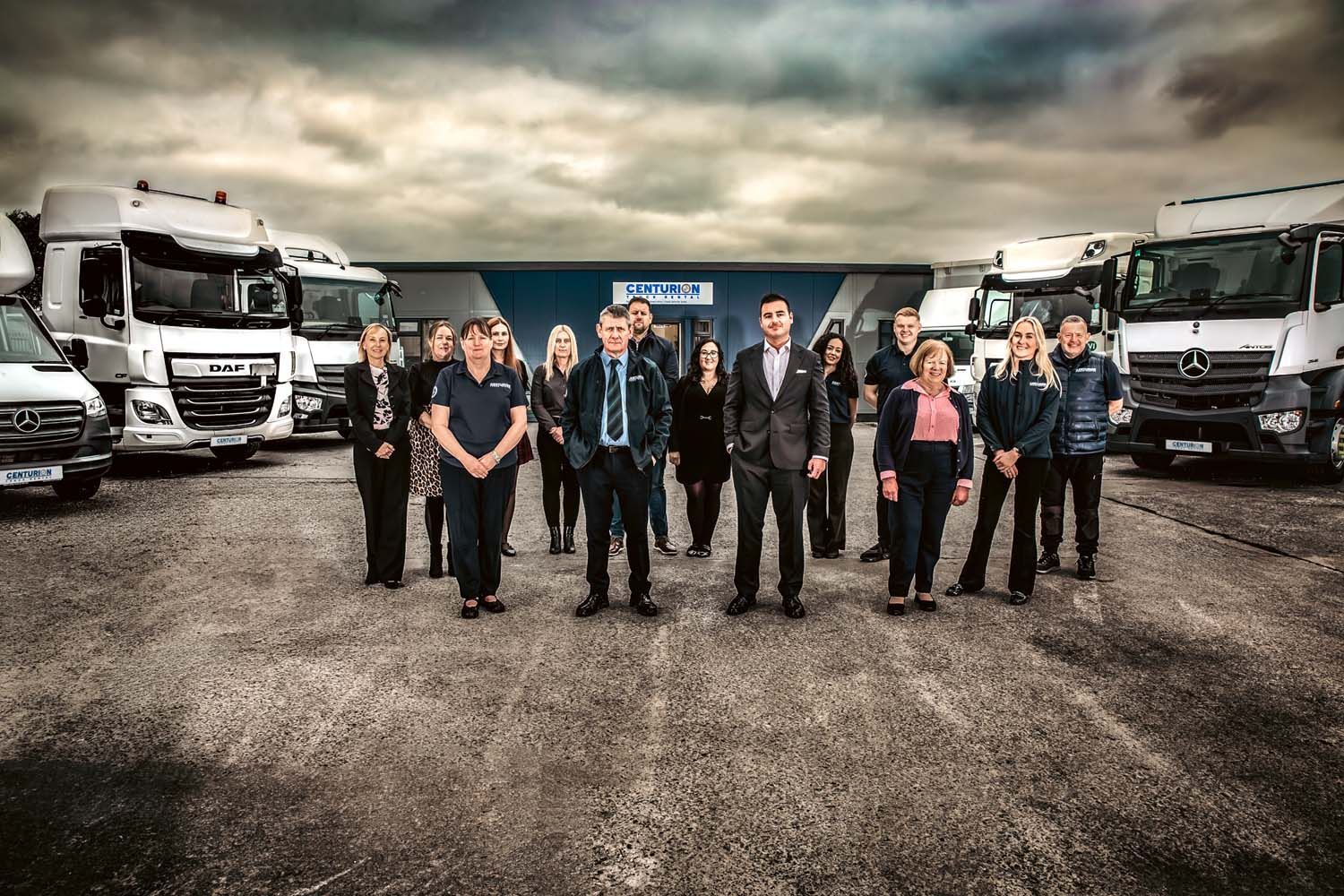 Truck fleet with a group of uniformed workers standing in a row on a gravel lot under a cloudy sky