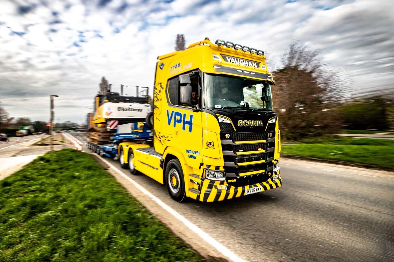 Yellow tow truck driving on a road, hauling a trailer under a cloudy sky