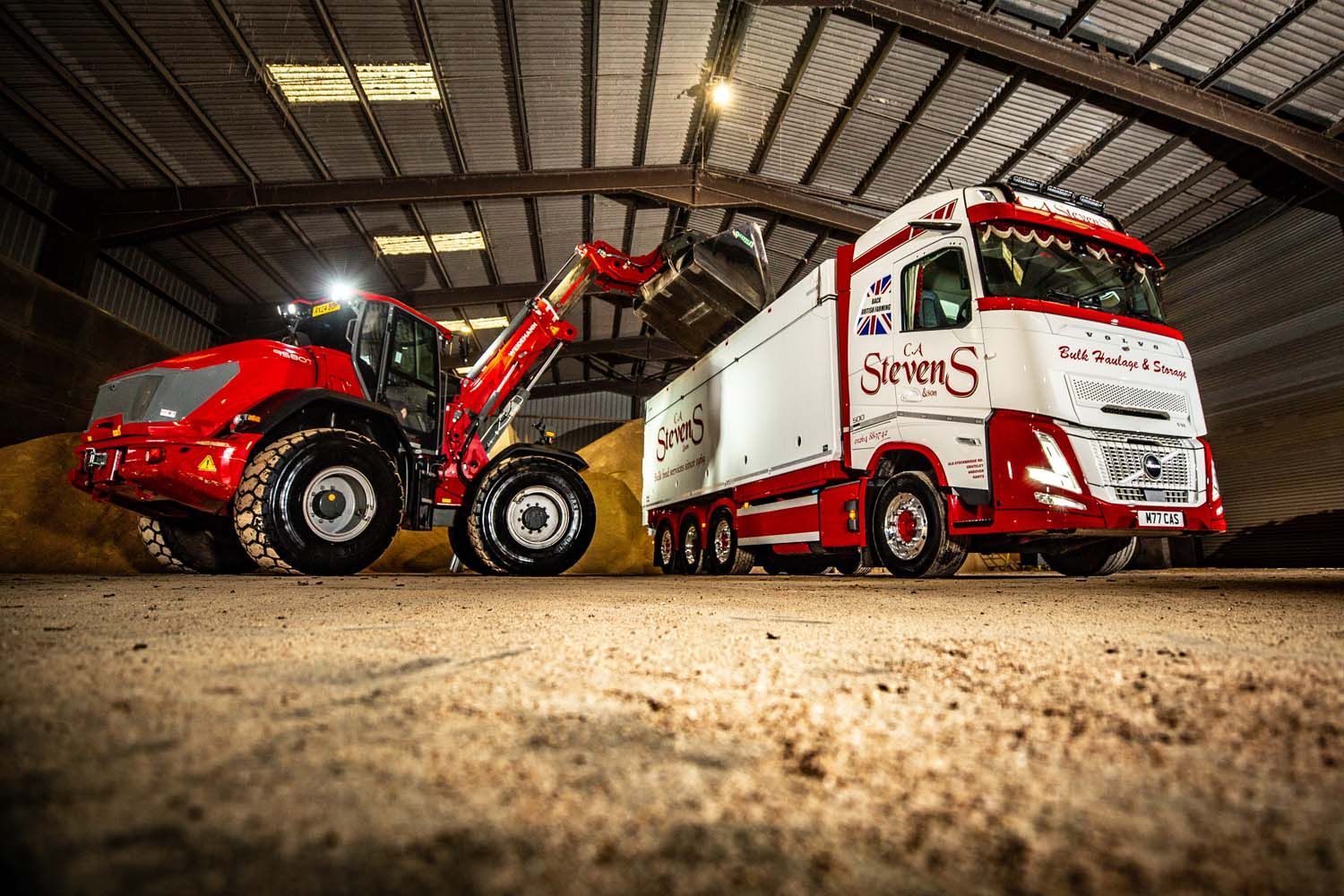 Commercial truck photography of red loader lifting a red-and-white trailer inside a warehouse, viewed from ground level