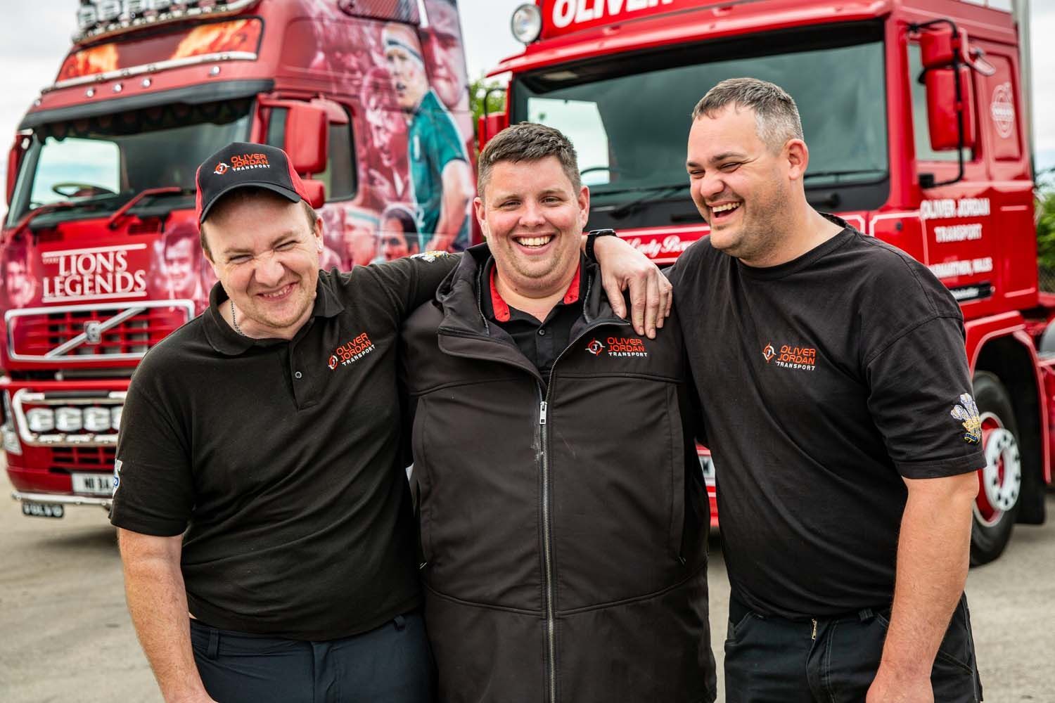 Three smiling men in black shirts standing in front of red trucks