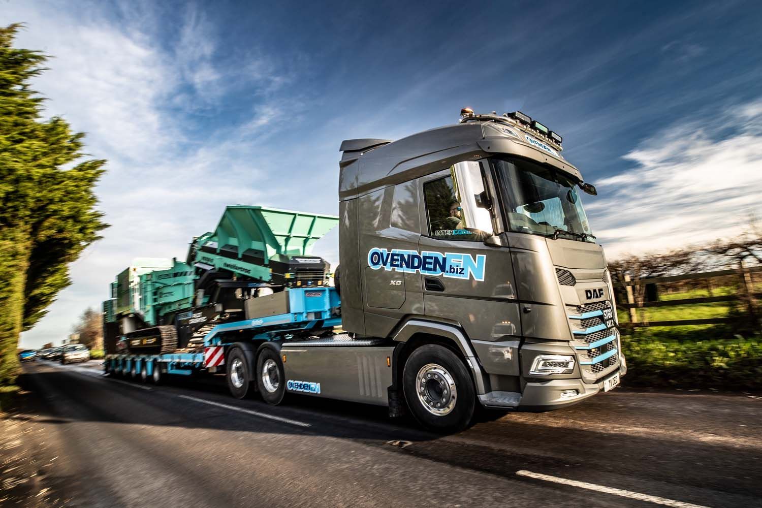 Commercial truck photography showing silver Mercedes truck hauling a green excavator on a flatbed trailer along a rural road