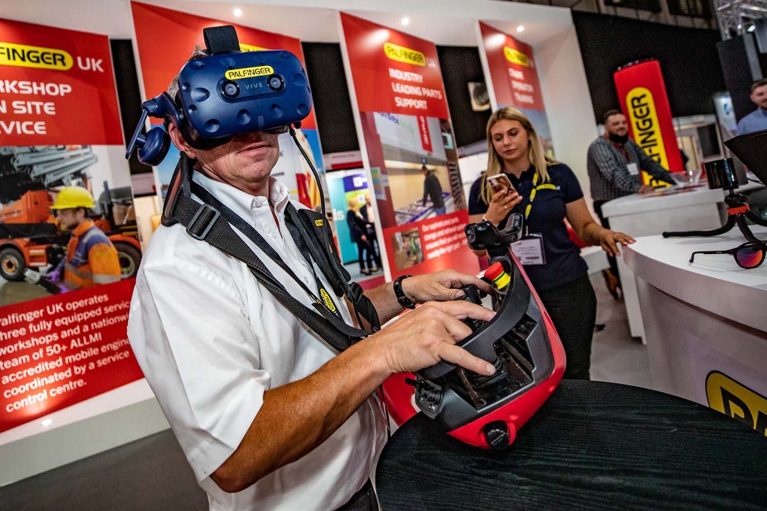 Person wearing a blue VR headset and holding controls at a trade-show booth with red signage