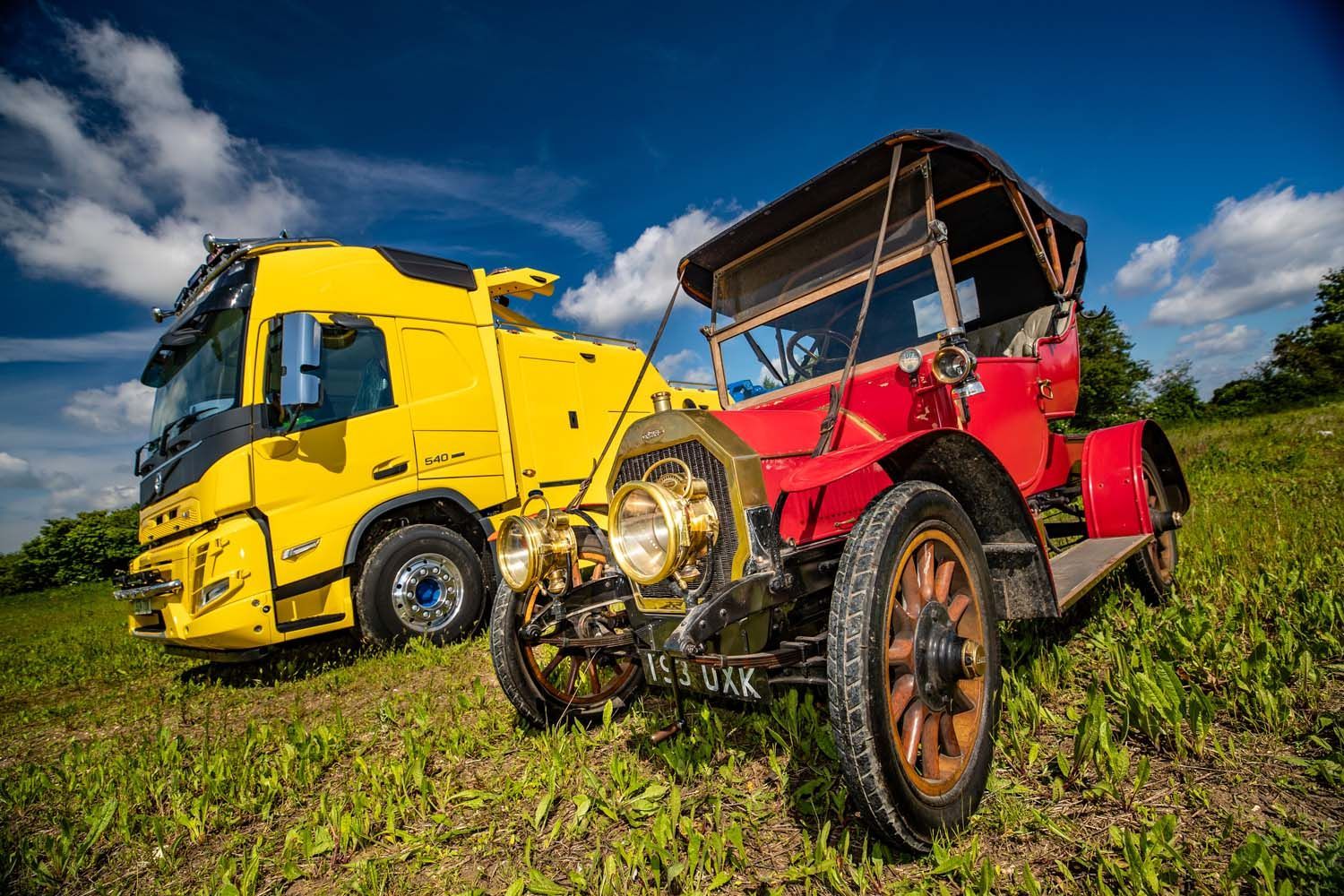 Yellow truck and red vintage off-road vehicle in a grassy field under a blue sky