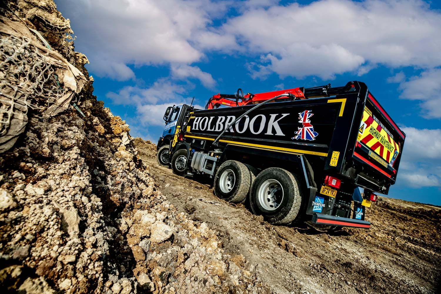 Black MOLBROOK dump truck driving up a rocky dirt slope under a cloudy sky