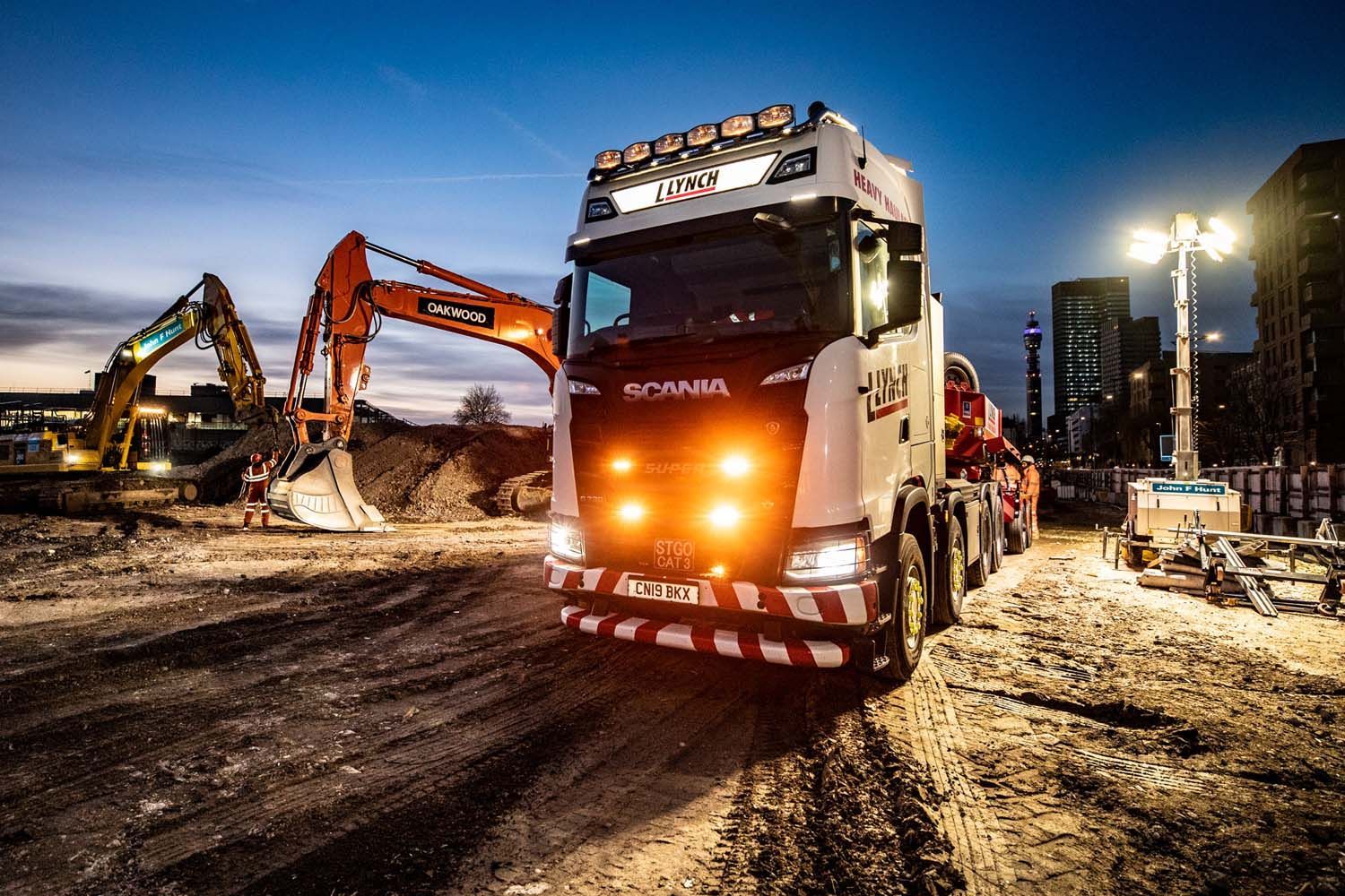 Heavy construction trucks and excavator at a muddy worksite at dusk, with bright headlights and floodlights