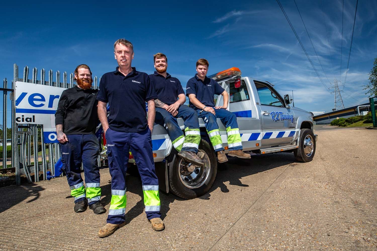 Four utility workers photographed in navy uniforms stand beside a white service truck on a sunny roadside.