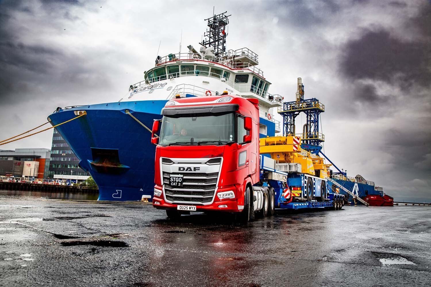 Red lorry parked in front of a large cargo ship at a wet shipping port under stormy skies