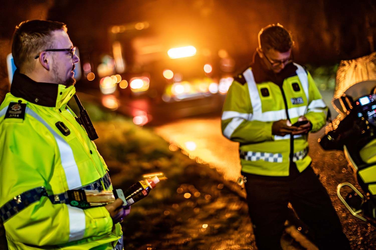 Two police officers in yellow jackets examining a roadside accident scene at night