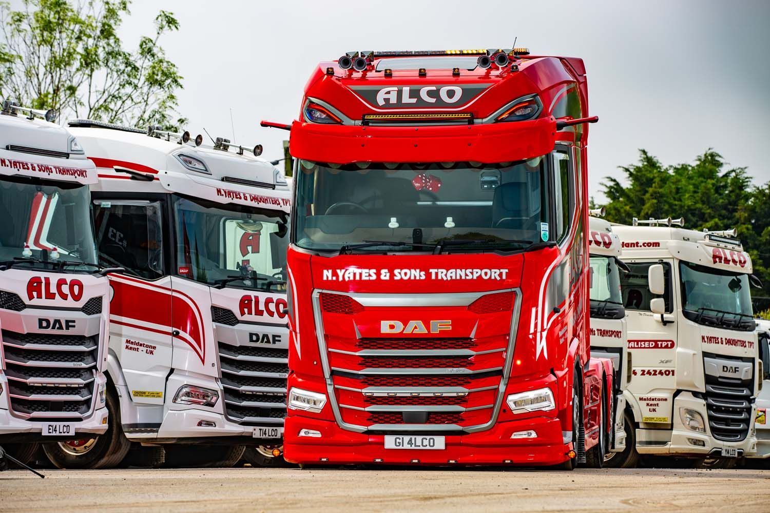 Truck photography of red ALCO cargo truck centered among white ALCO trucks in a row on a lot
