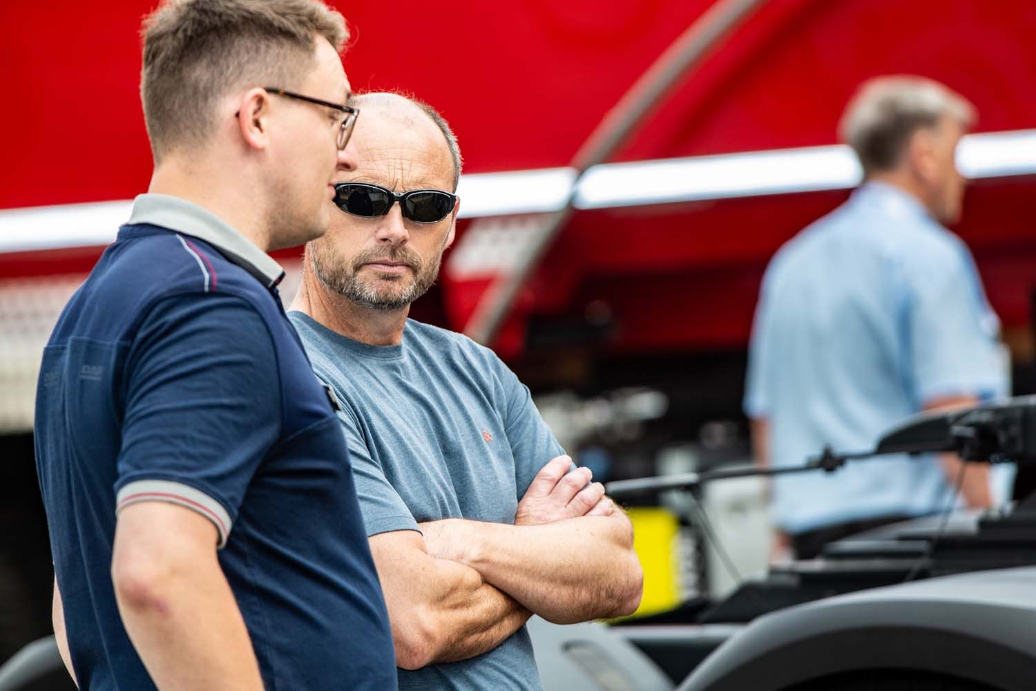 Two men talking beside a red car, one wearing sunglasses with arms crossed