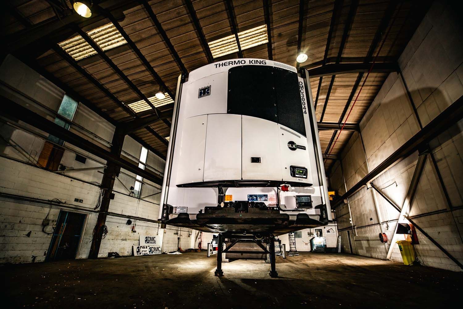 Large white vehicle on a lift inside an industrial garage, with workers nearby.