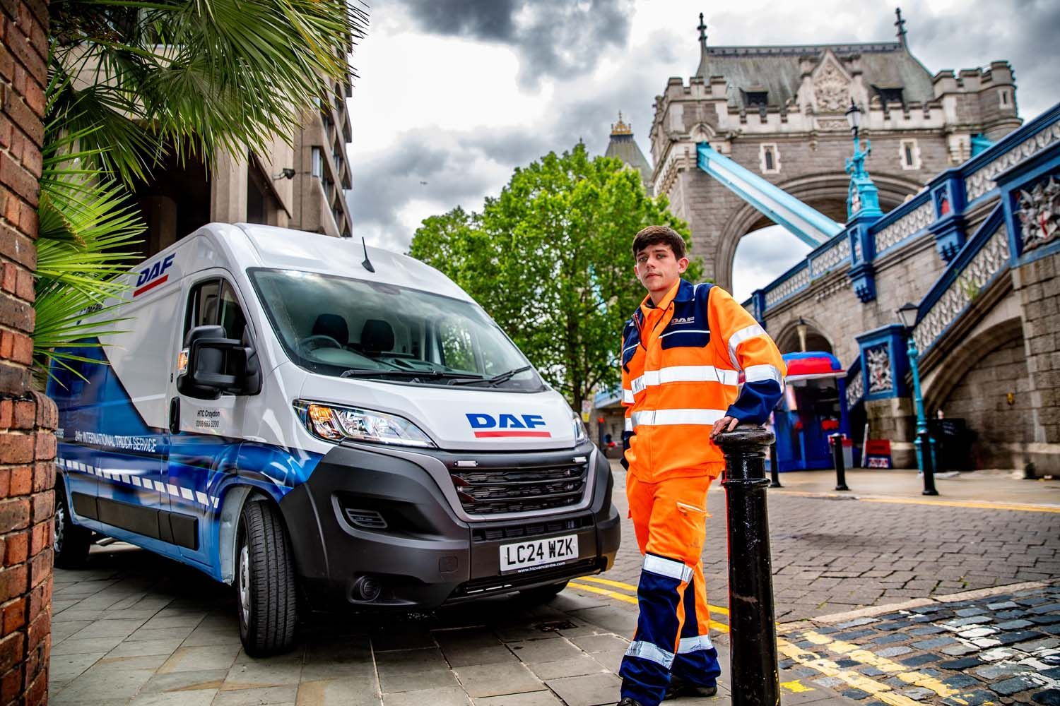 Worker in orange hi-vis stands beside a blue service van on a cobblestone street near a stone bridge.