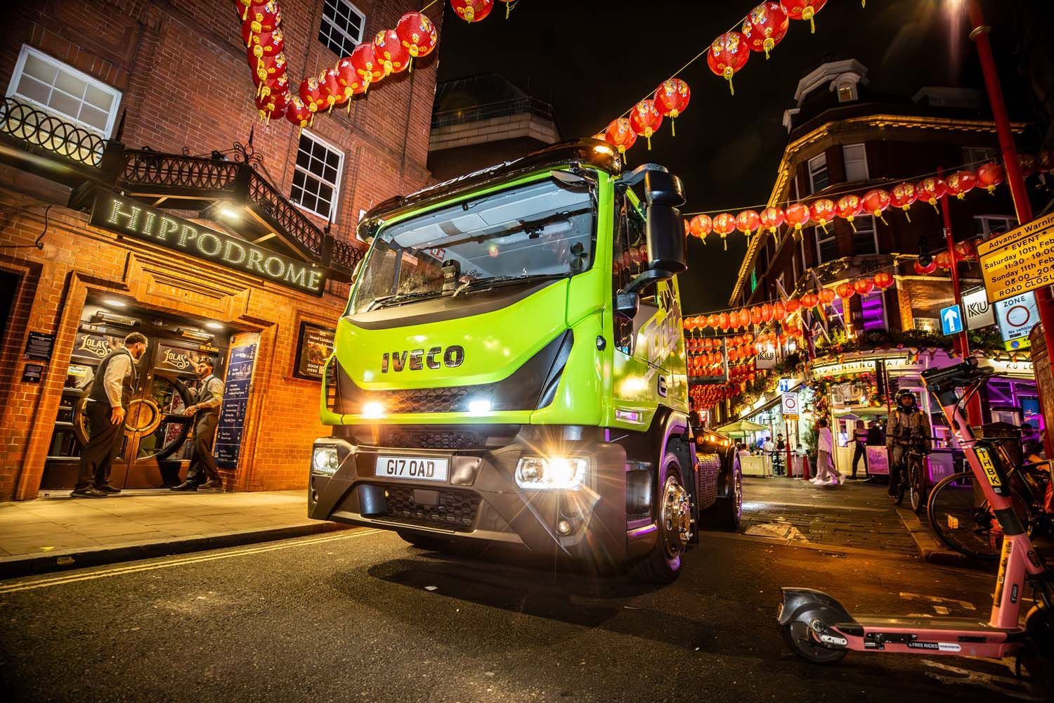 Green truck photographed driving down a wet, lantern-lit street at night in a bustling Chinatown