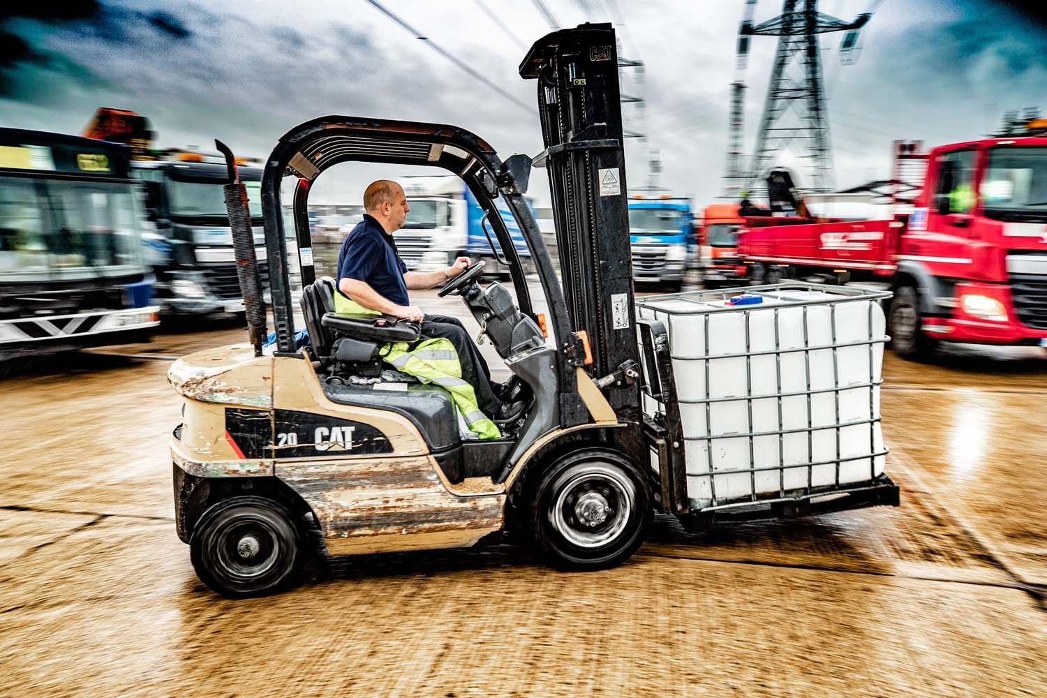 Forklift carrying a white tank across a muddy worksite, with trucks and cranes in the background