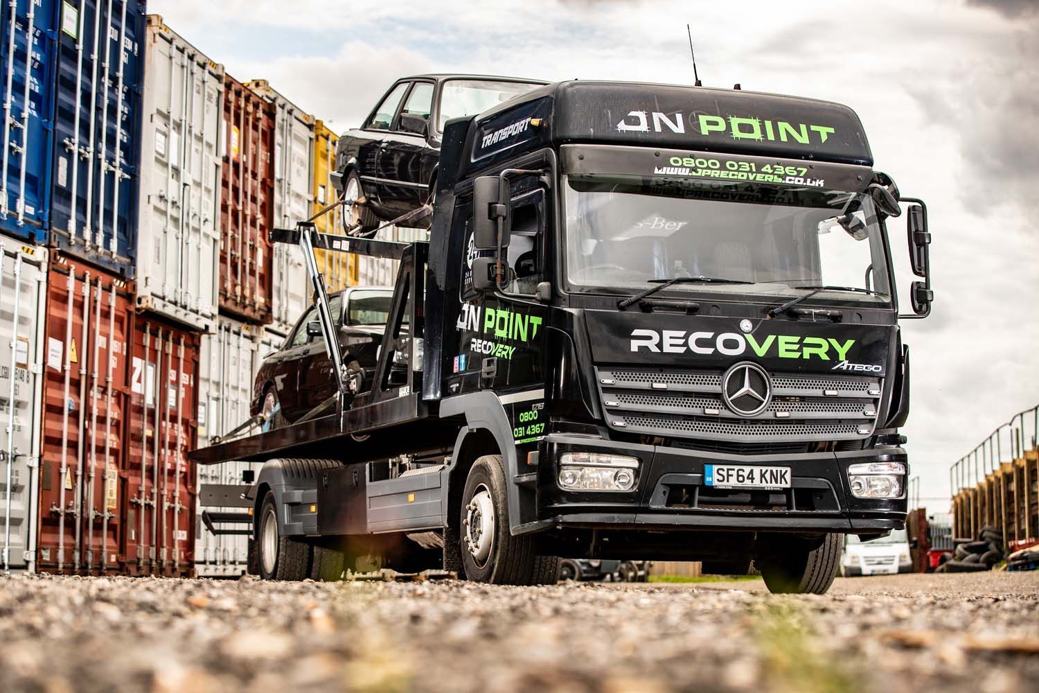 Black recovery truck beside stacked shipping containers in an industrial yard