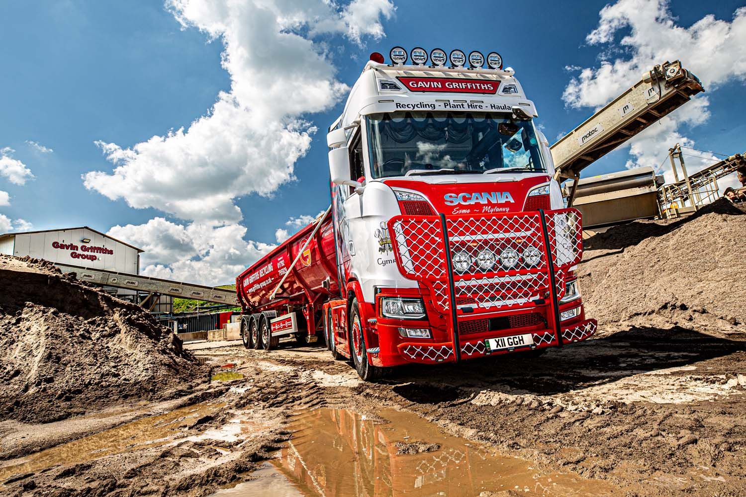 Red dump truck hauling mud at a muddy construction site under a cloudy sky
