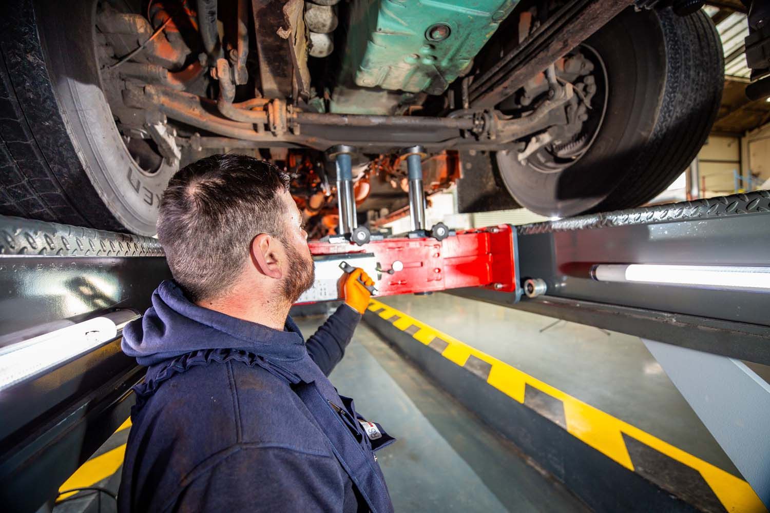 Photograph of truck mechanic inspecting a lorry’s undercarriage on a lift in a garage with yellow-and-black safety markings