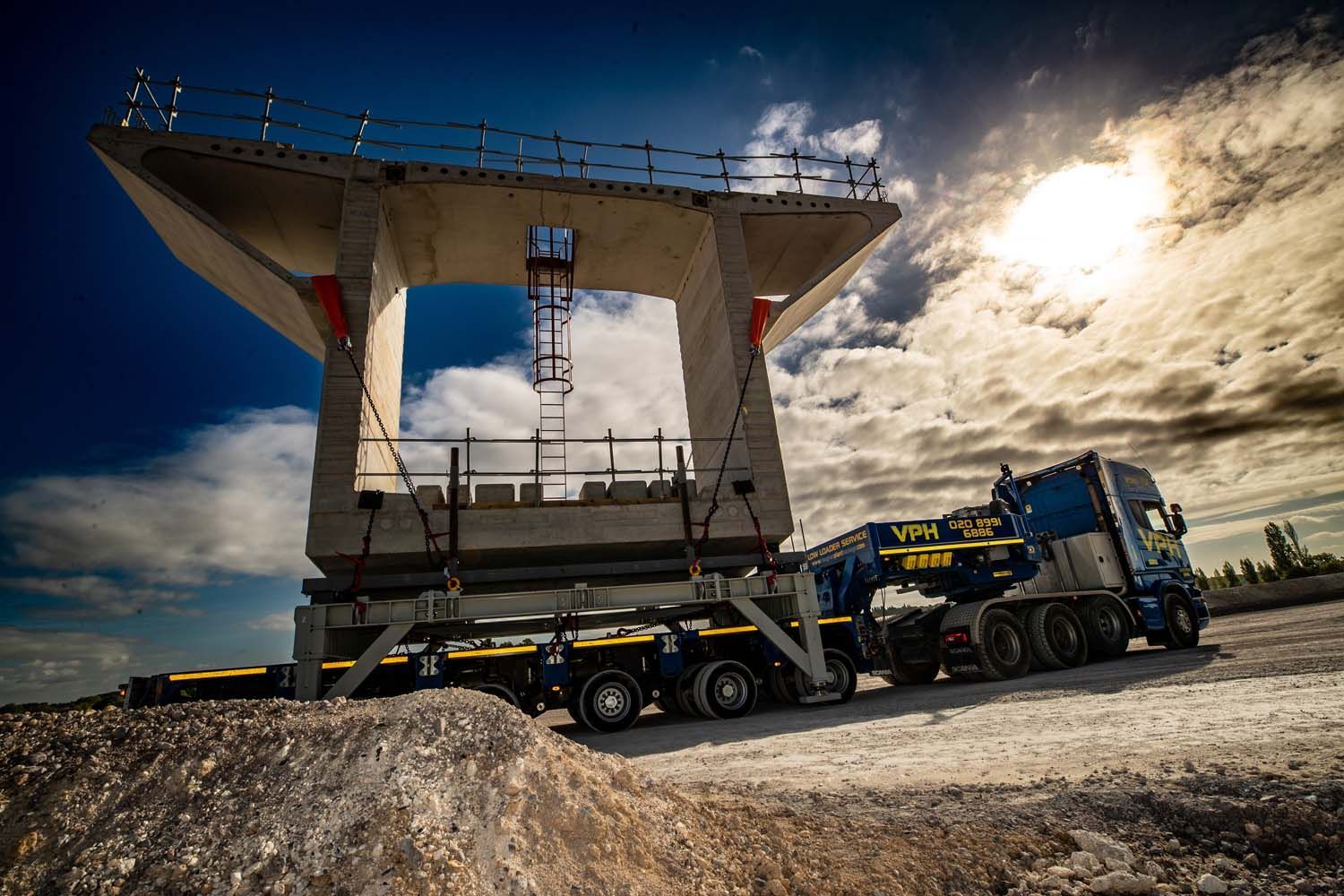 Heavy mining haul truck under a massive dump hopper, backlit by bright sun at a quarry.