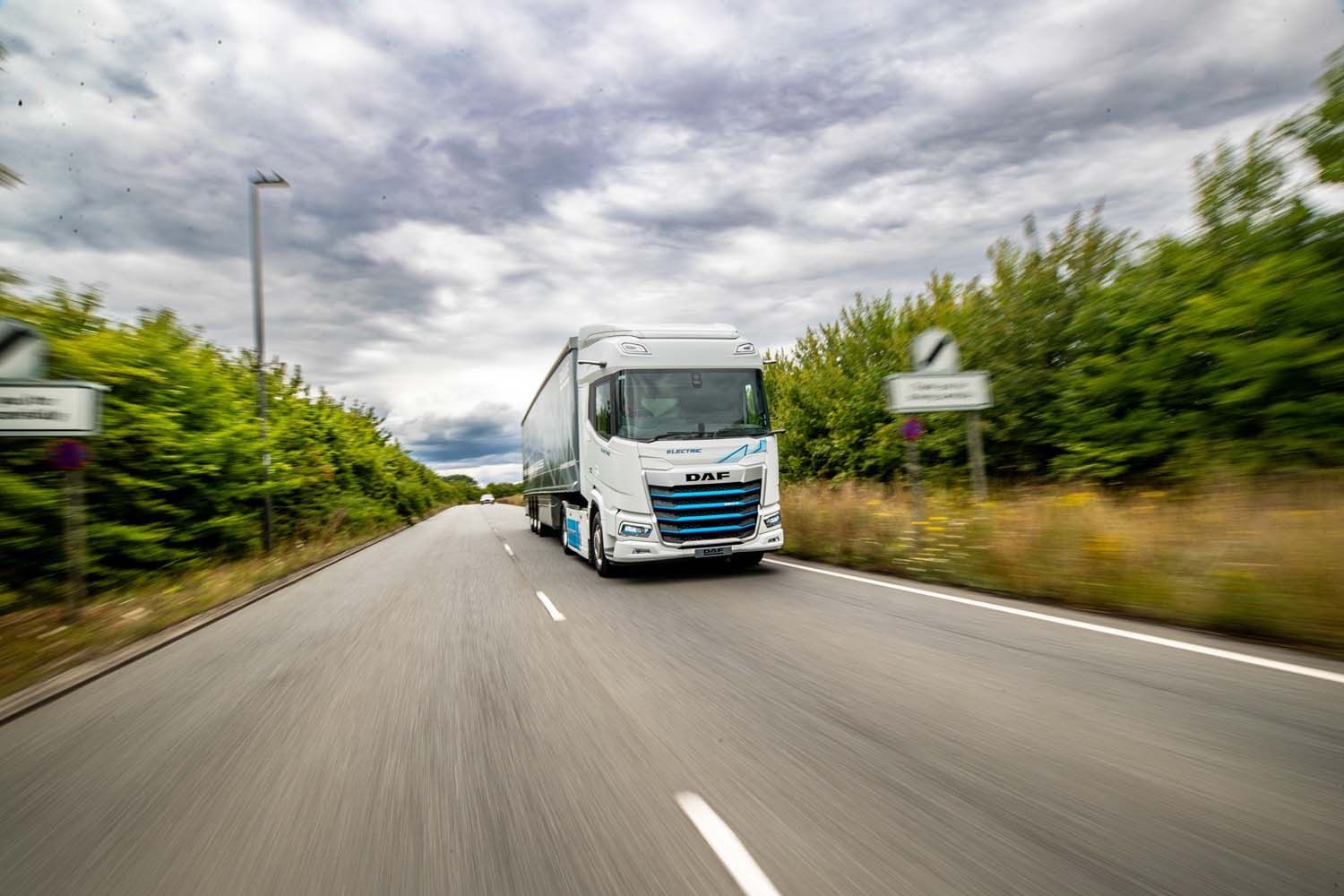 White semi-truck driving on a two-lane road beside green hedges under cloudy sky