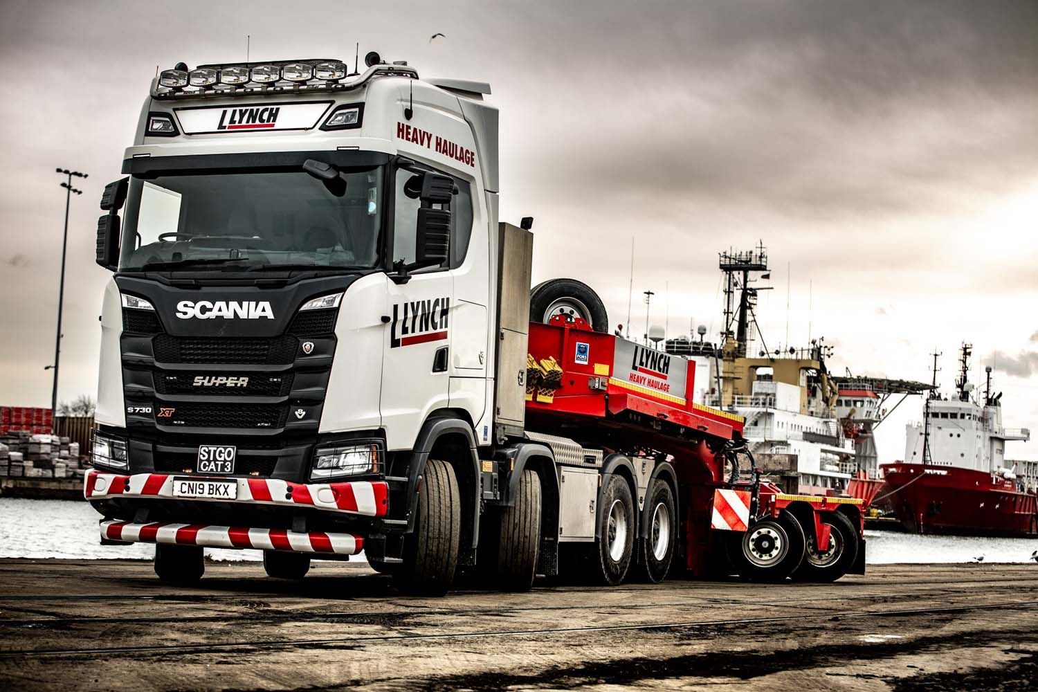 Commercial truck photography red Scania heavy-haul truck with lowboy trailer and excavator on a roadside under cloudy skies