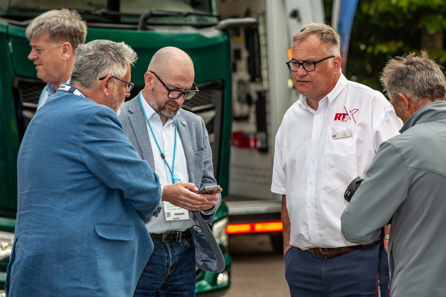 Five men stand talking outdoors near a green truck, one holding a phone and another wearing a white shirt.