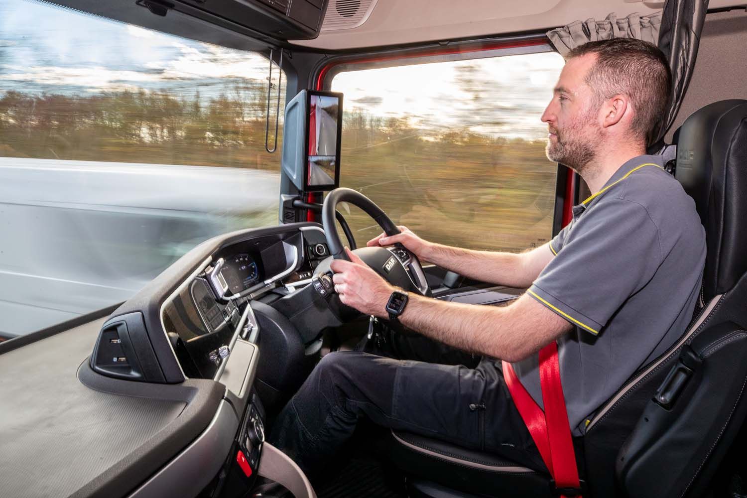 Driver steering a truck on a road, viewed from the cab with blurred scenery outside.