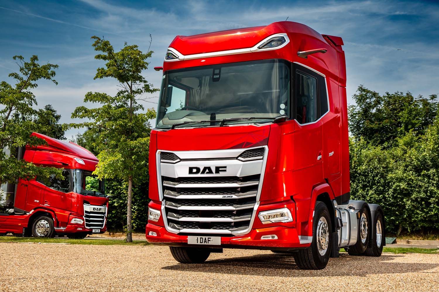 Bright red DAF semi-truck parked on a gravel lot, with another red truck in the background and trees behind.