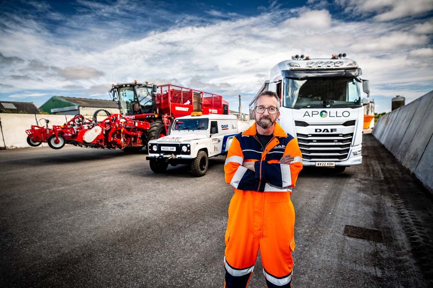 Worker in orange safety gear standing in front of trucks and red equipment in an industrial yard