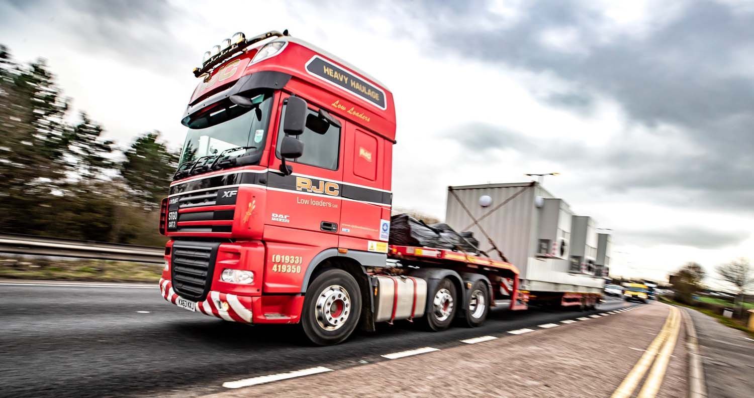 Red lorry hauling a flatbed trailer on a wet road near industrial buildings