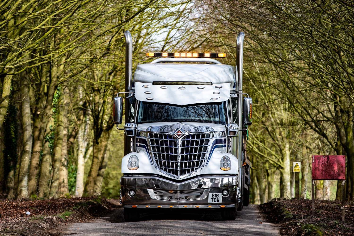 White semi-truck driving down a tree-lined road, front view with headlights on