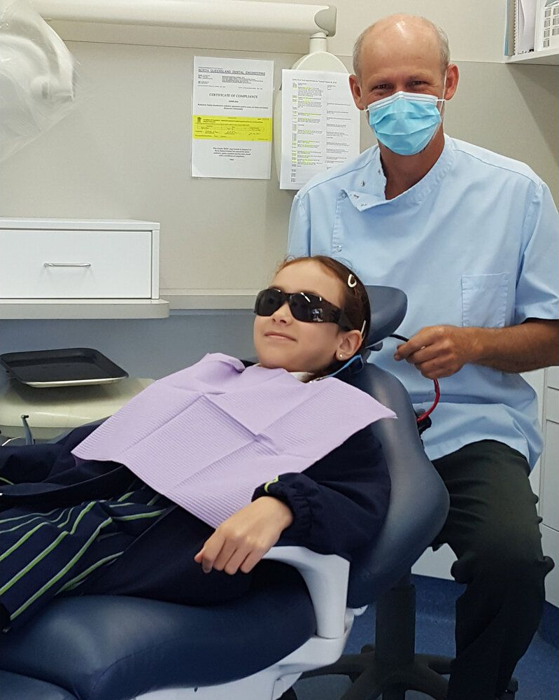 A dentist wearing a mask stands behind a patient in a chair wearing protective eyewear in a dental office.