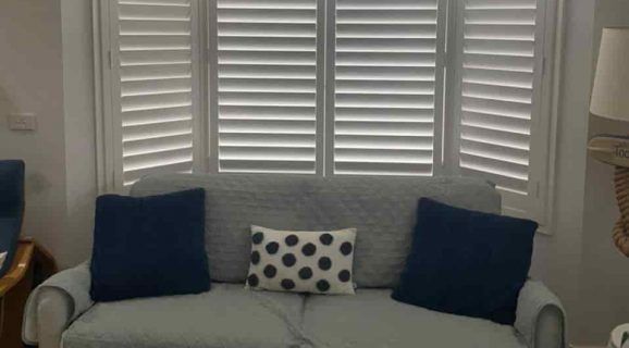A Balcony with A Couch and Pillows and Shutters on The Windows — Bay Blinds & Doors in Lake Munmorah, NSW