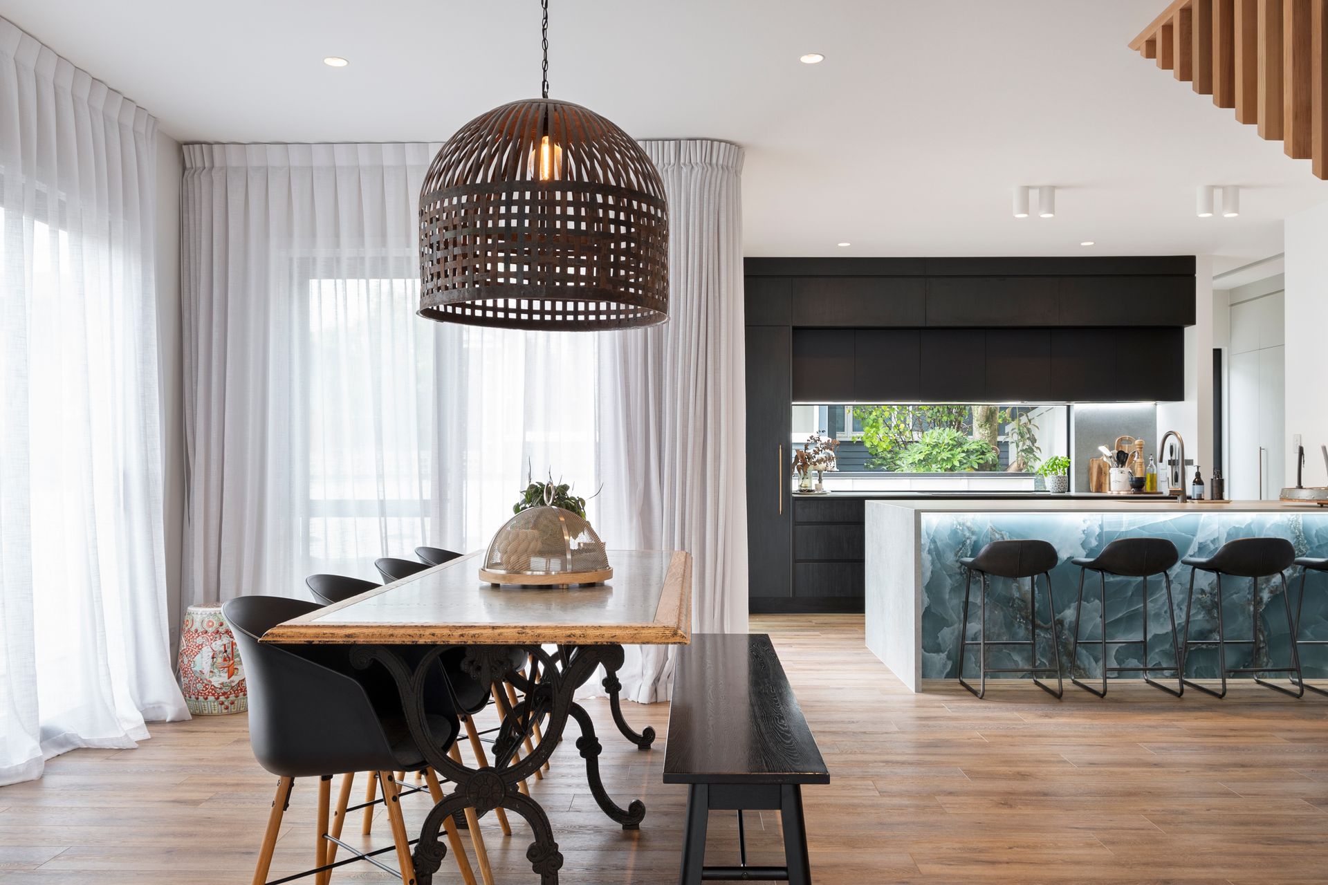 Modern open-plan dining and kitchen area with a wood table, black chairs, woven pendant light, and blue island stools