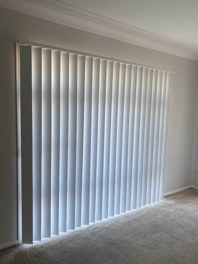 A Living Room with White Vertical Blinds on The Window — Bay Blinds & Doors in Bateau Bay, NSW
