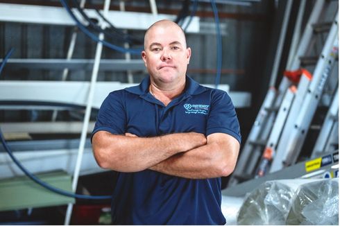Man standing with arms crossed in a workshop, wearing a navy polo shirt.