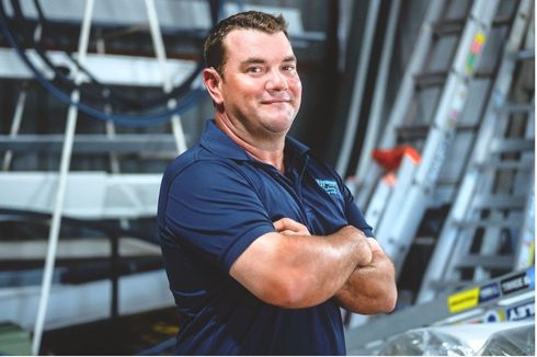 Smiling man in a navy polo stands with arms crossed in an industrial setting