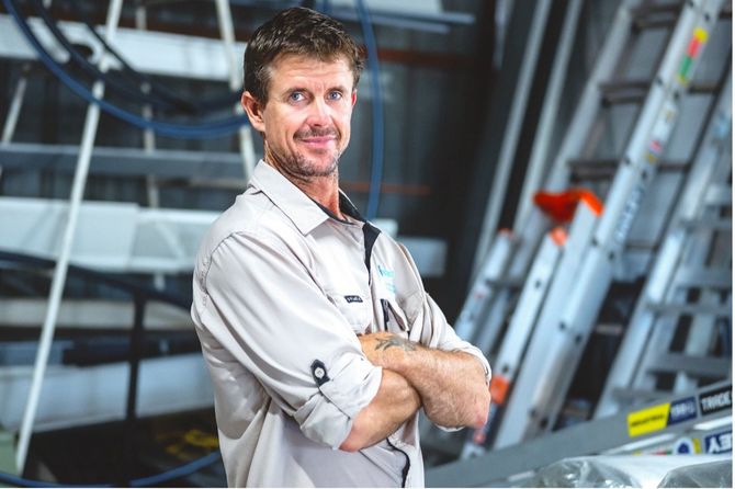 Man standing with arms crossed in an industrial workshop, wearing a light shirt, with ladders and machinery behind him