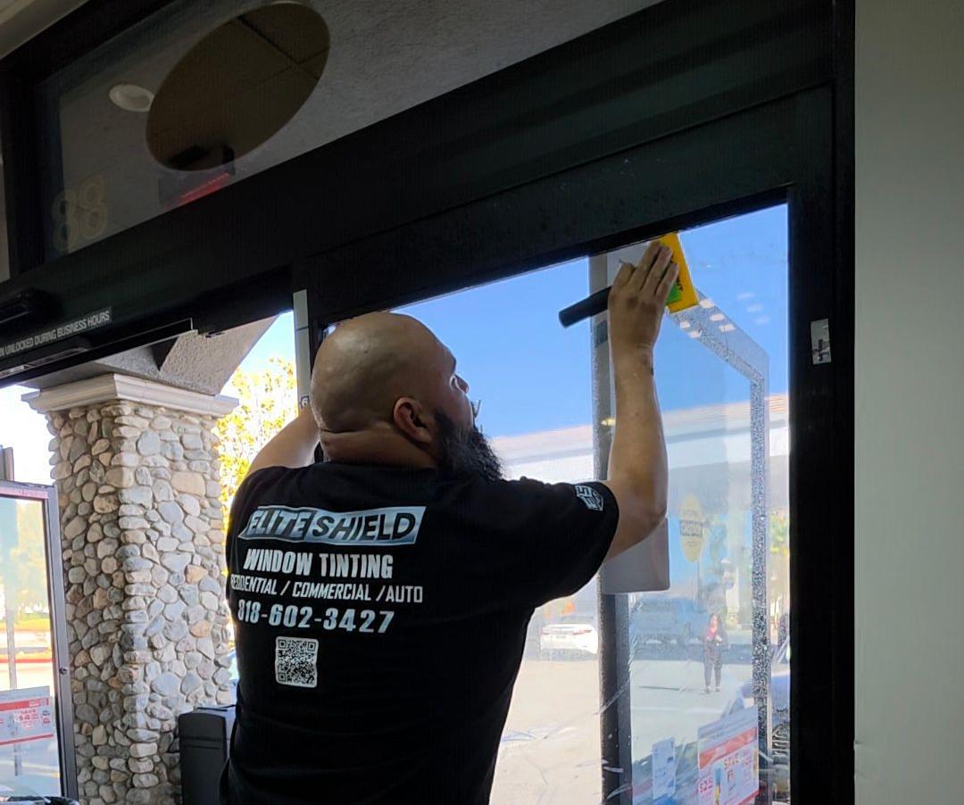 Man installing window tint in a store, using a squeegee. He wears a black shirt with business logo.