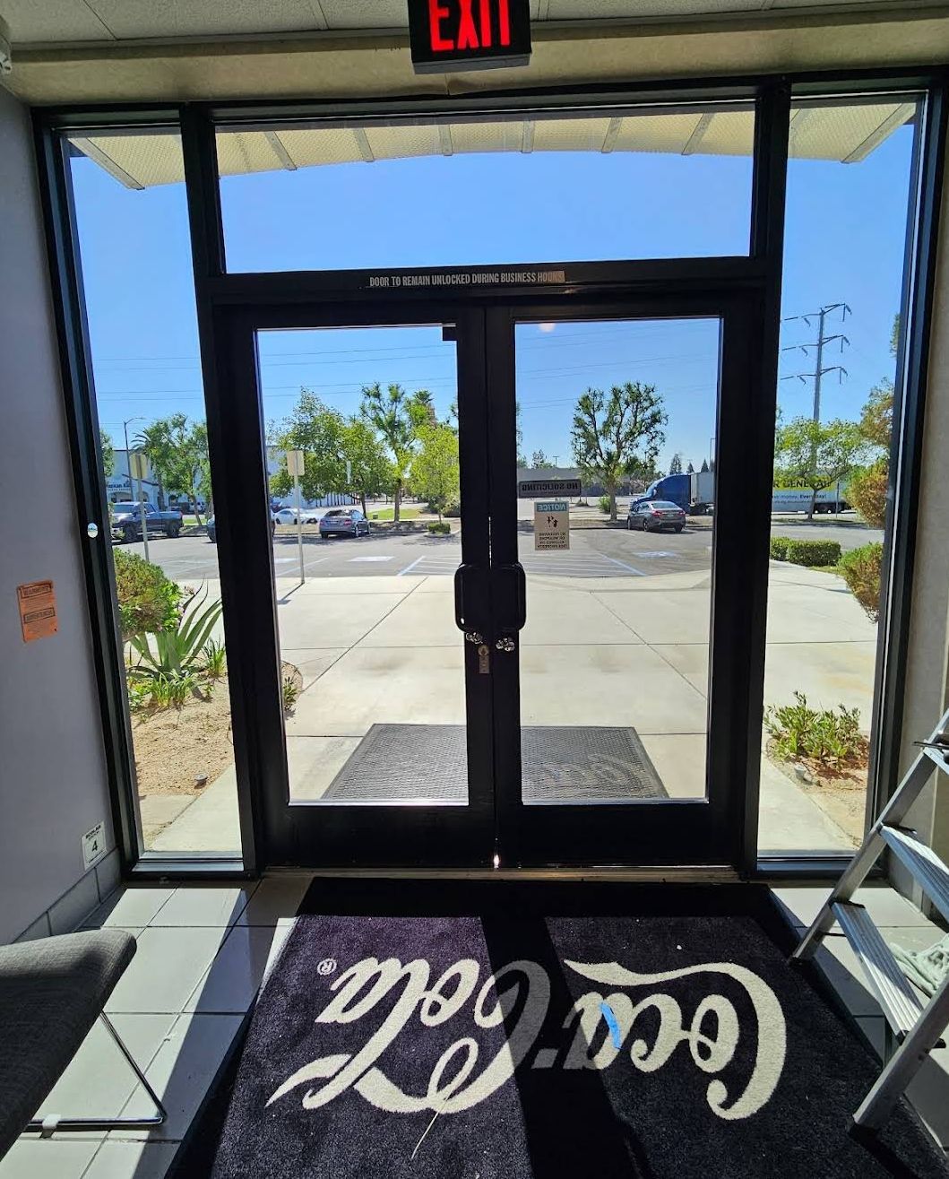 Store entrance with glass doors, EXIT sign, and Coca-Cola mat facing a sunny parking lot