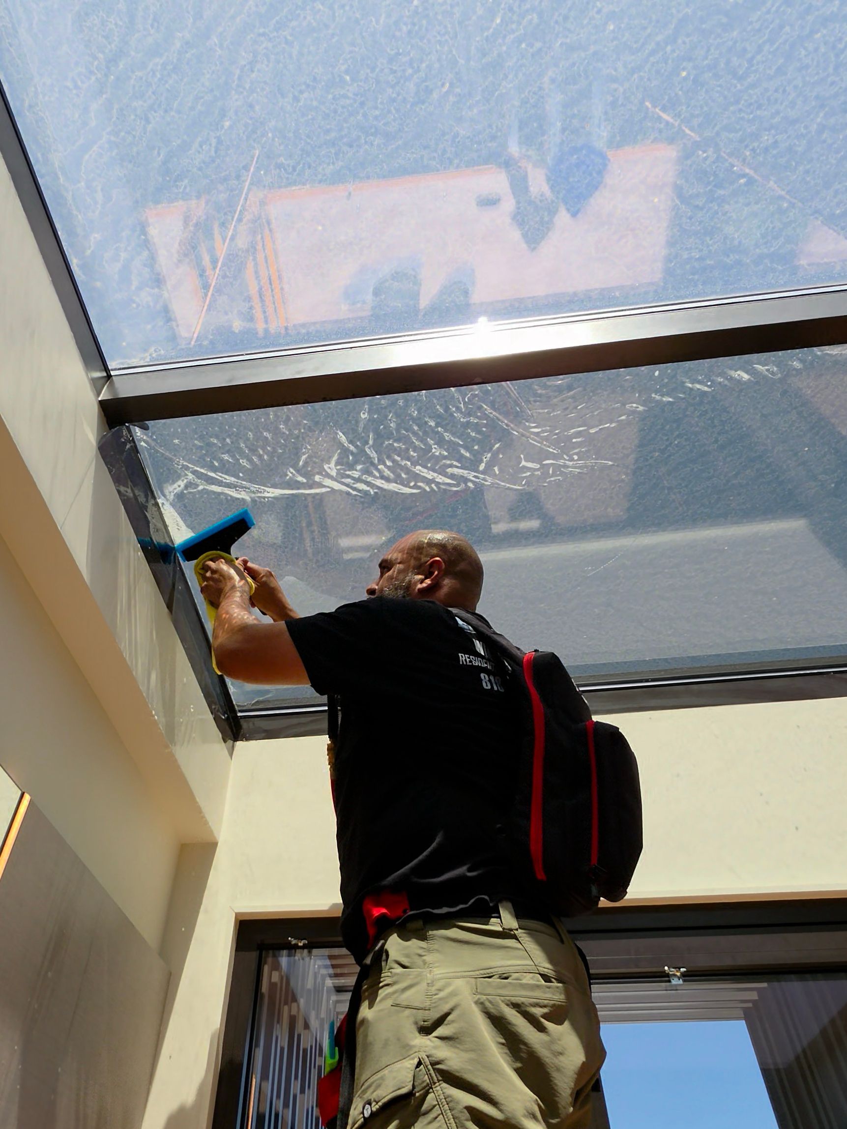 Person cleaning a skylight window indoors with a blue cloth, wearing a black shirt and beige pants.