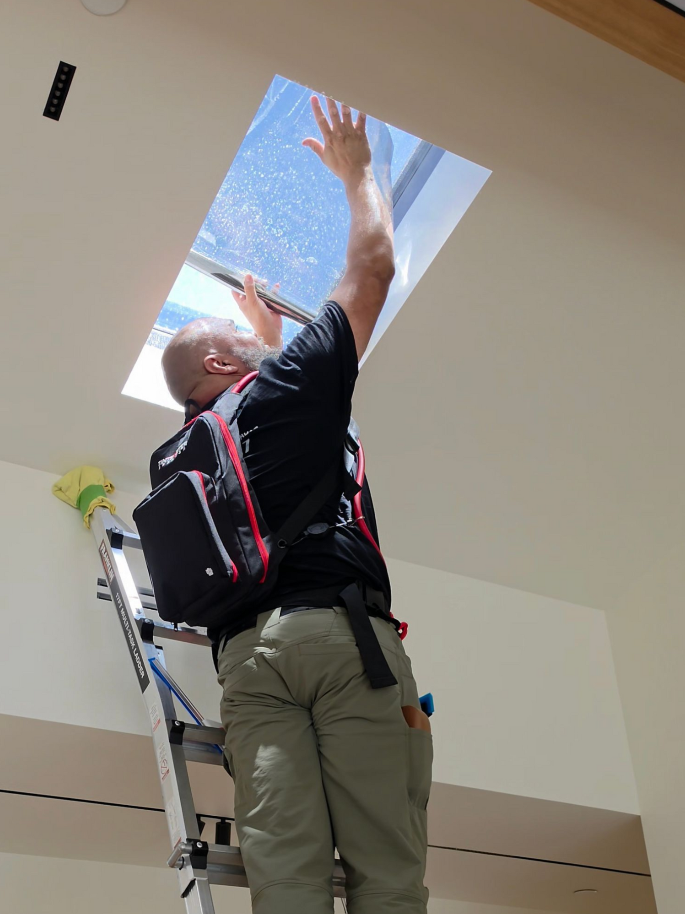Worker reaching up through an open ceiling panel on a ladder indoors.