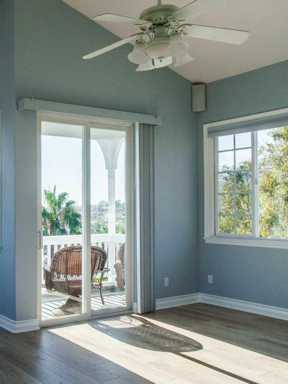 Empty blue bedroom with ceiling fan, sliding glass door, and sunny windows overlooking a patio.