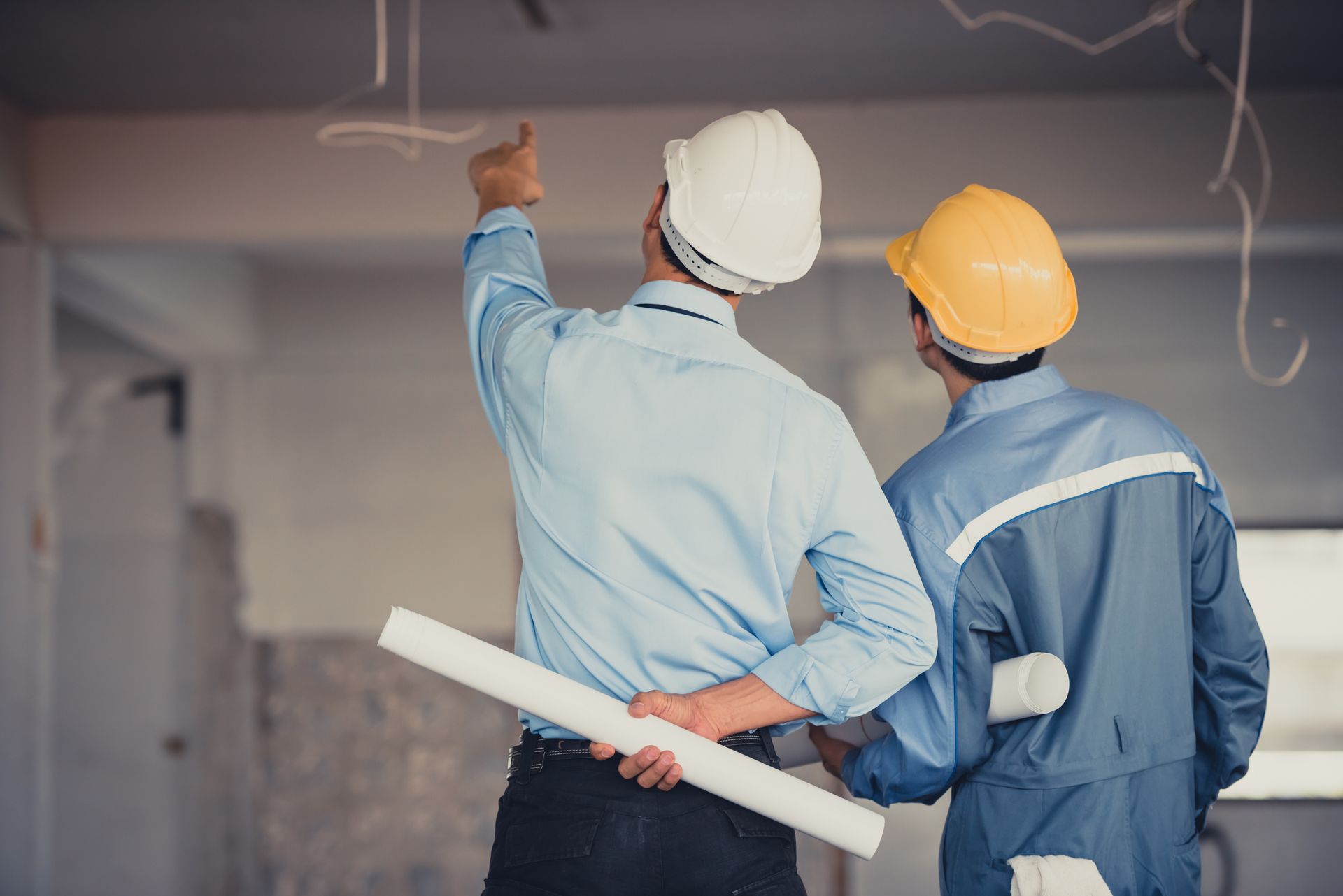 Two construction workers in hard hats reviewing plans and pointing at ceiling wiring.