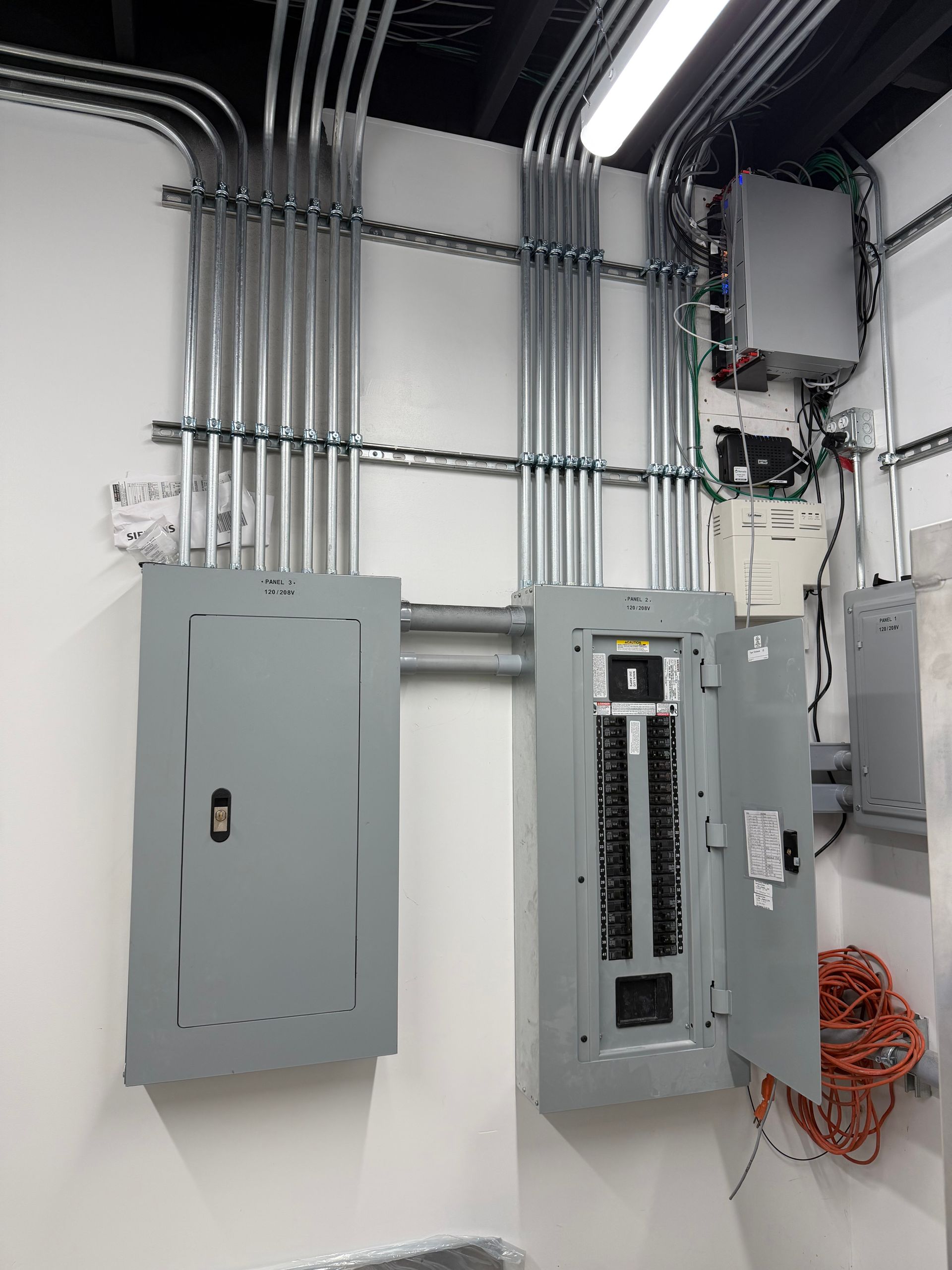 Two grey electrical service panels mounted on a white wall, connected by extensive metal conduit piping overhead. Two grey electrical service panels mounted on a white wall, connected by extensive metal conduit piping overhead.