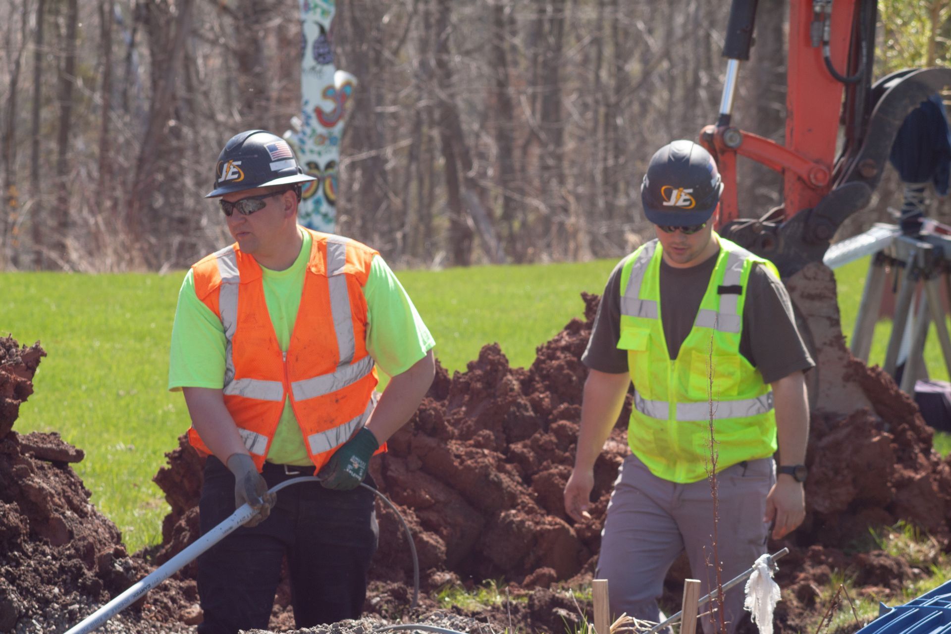 A yellow excavator with a person in the cab sits in a field near a red and white traffic cone.