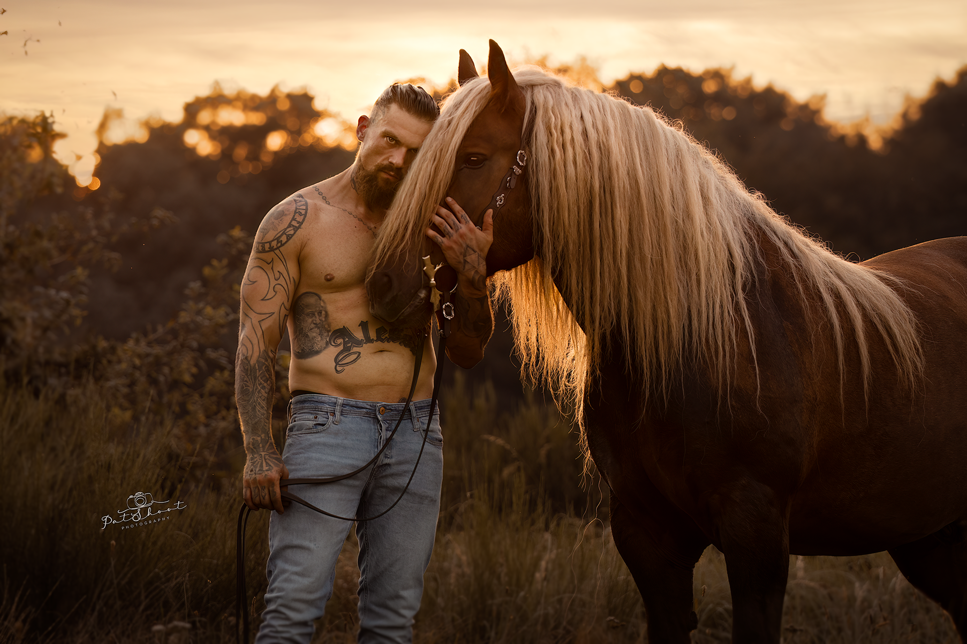 Een man met tatoeages aait een bruin paard met een lange blonde manen bij zonsondergang.
