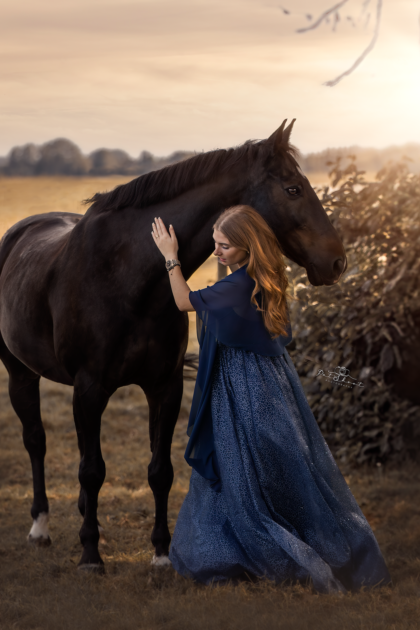 Vrouw in een blauwe jurk aait een donker paard in een veld bij zonsondergang.