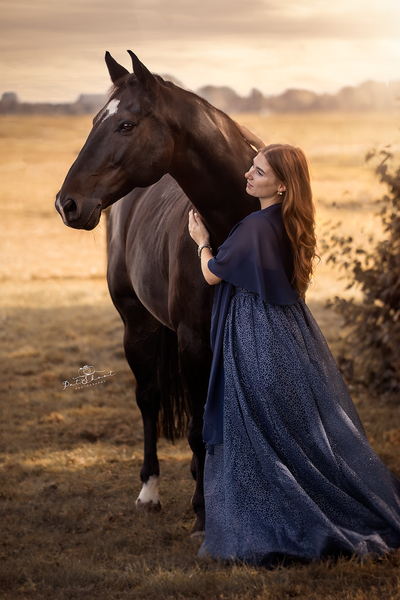 Vrouw in een glinsterende blauwe jurk aait een donker paard in een veld bij zonsondergang.