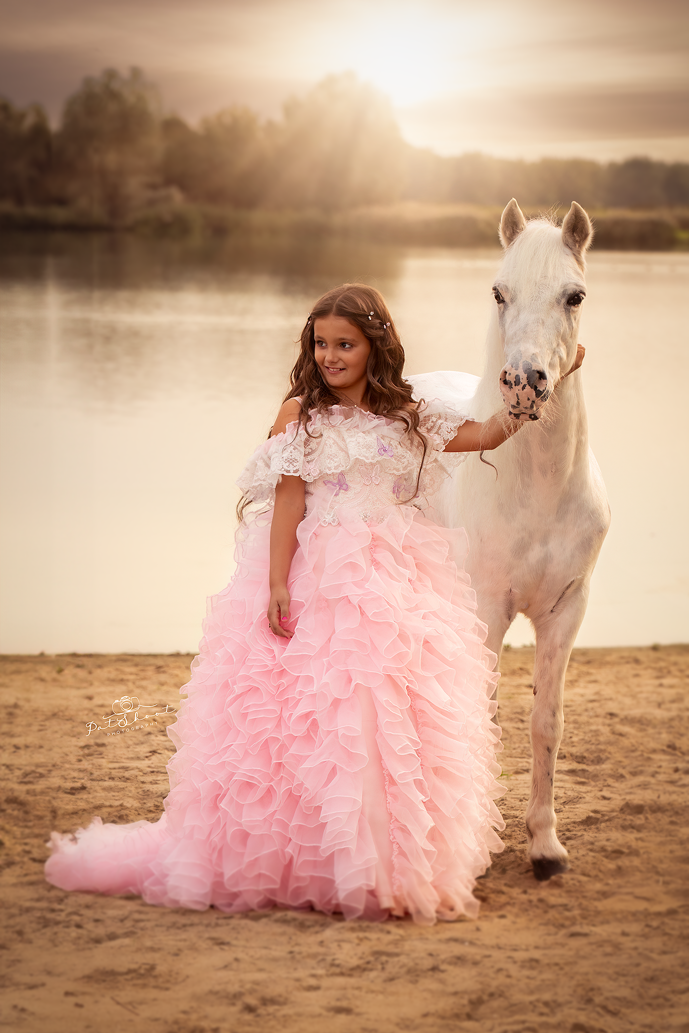 Meisje in een roze jurk met een wit paard bij een meer, beiden verlicht door de zon.