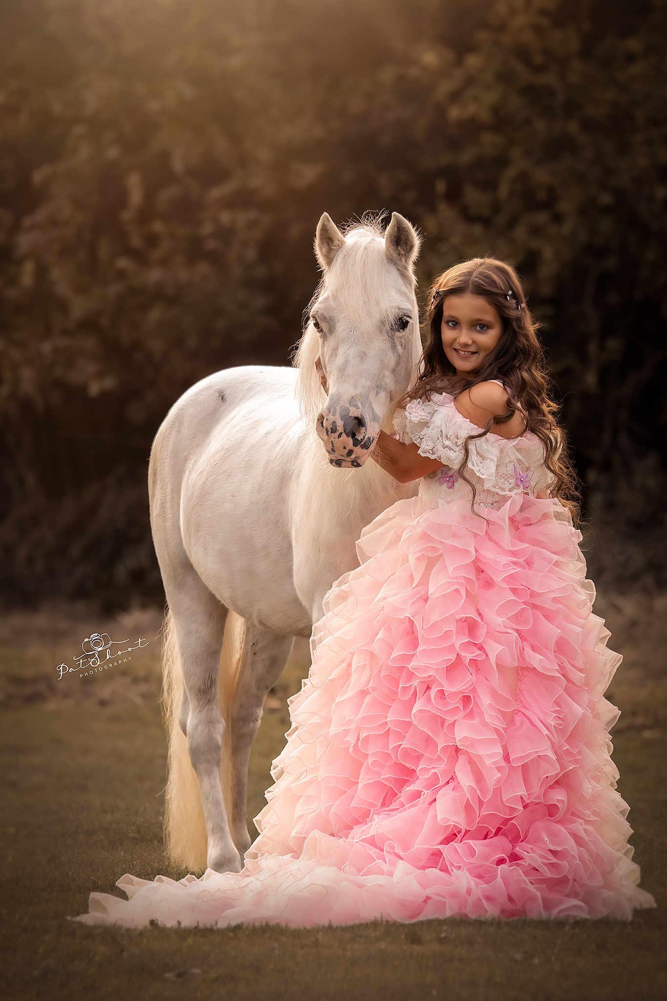 Een meisje in een roze jurk met ruches omhelst een witte pony in een veld.